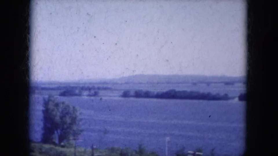 1969 8mm home-movie frame showing a broad lake view in Lake Canyon, Arizona, with distant trees, low hills, and pale sky.