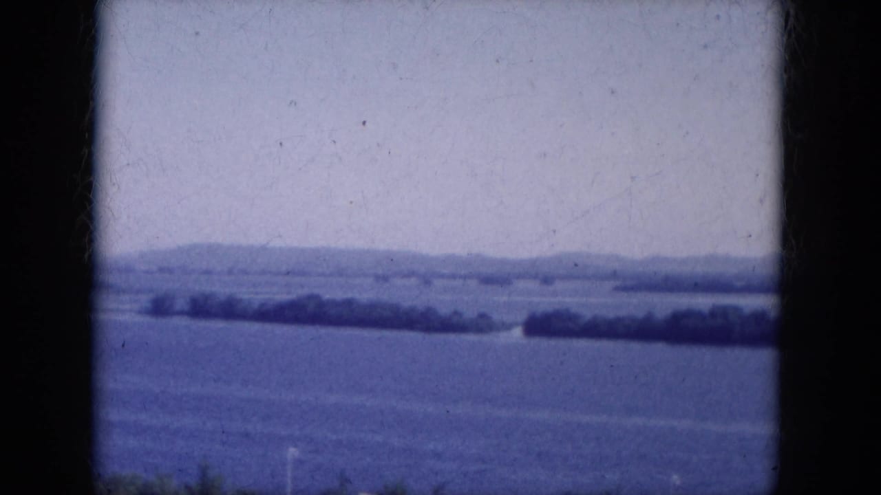 Static waterscape with distant shore, low hills, pale sky, and dark film borders.