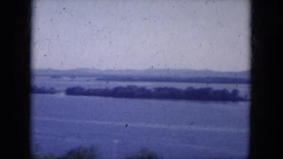Blue-tinted 1969 8mm frame showing Lake Canyon water, a tree-lined island or shore strip, and low hills in the distance.
