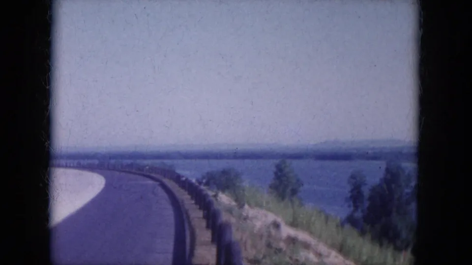 1969 8mm frame of a curving road along Lake Canyon, Arizona, with guardrail, shoreline brush, blue water, trees, and a hazy horizon.