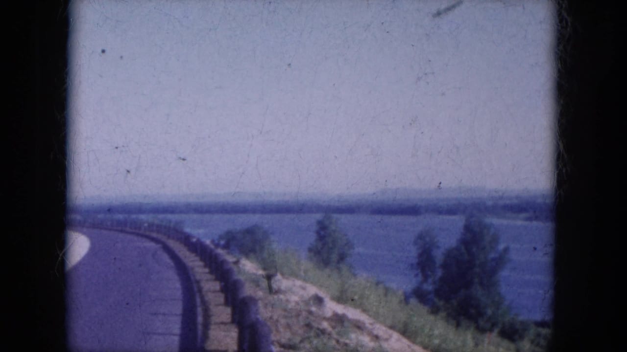 Curving roadside beside a broad blue river with trees and distant hills.