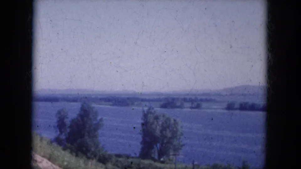 1969 Arizona travel footage frame showing blue water, two foreground trees, brushy bank, and low hills in the distance.