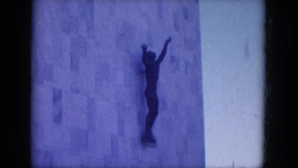 Close 1969 8mm view of the facade sculpture, a dark figure with one arm raised against a pale stone wall in Arizona.