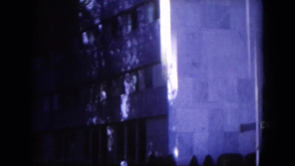 Blue-tinted 8mm frame showing a darker windowed section beside a pale exterior wall, with a small silhouetted person near the bottom right.