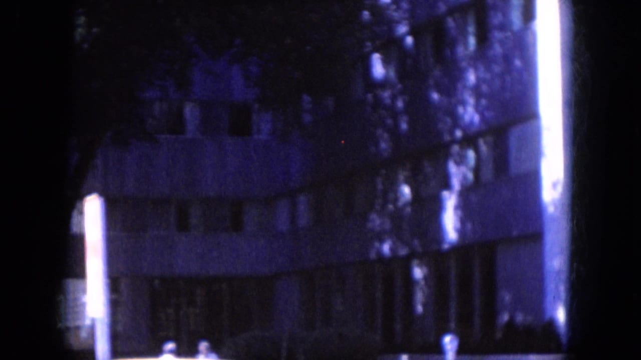 Blue-tinted view of a multi-story building partly hidden by trees and a bright vertical post.