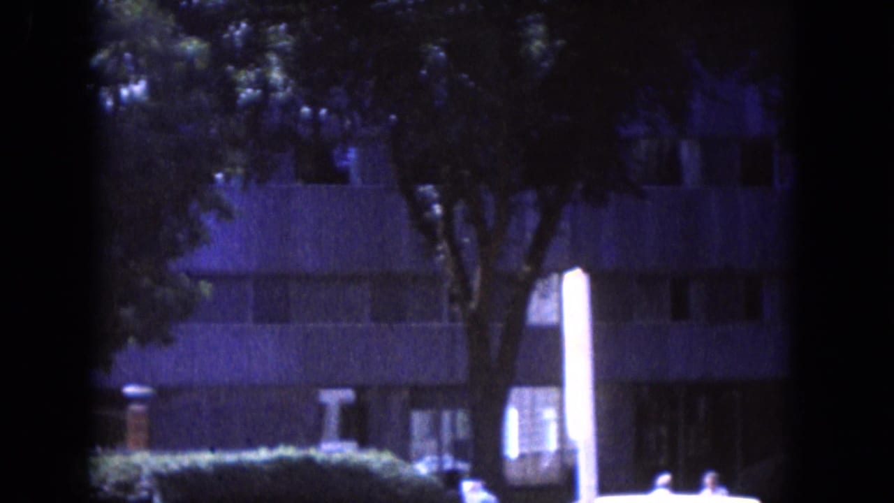 1969 Lake Canyon, Arizona frame with a blocky building facade, large tree trunk, shrubs, and bright vertical posts in blue-tinted 8mm color.