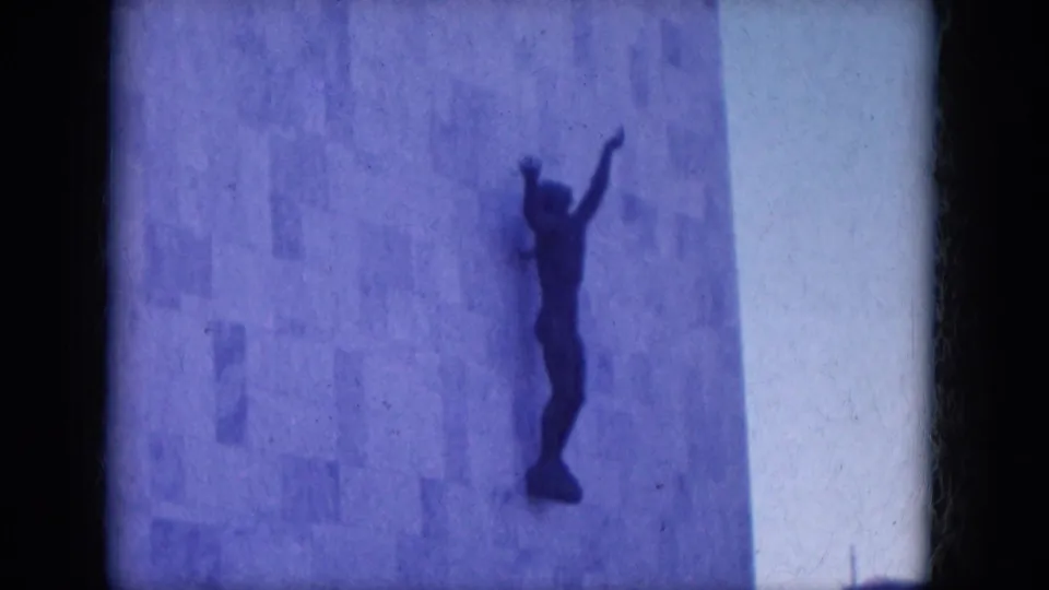 1969 Lake Canyon, Arizona 8mm frame of a dark raised-arm sculpture attached to a textured concrete wall.