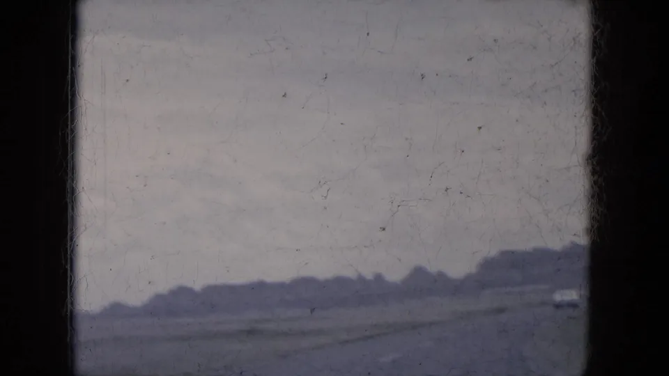 1969 8mm view from a moving vehicle in Lake Canyon, Arizona, with open roadway, low hills, gray sky, and a small vehicle in the distance.