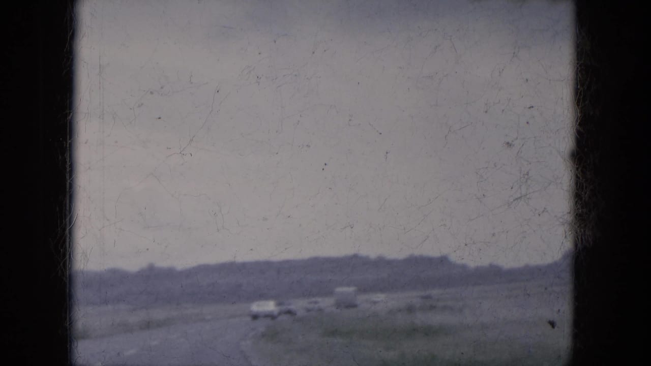 Passenger-view 8mm frame of a rural Lake Canyon road, with small vehicles ahead, sparse vegetation, and a low horizon beneath gray sky.