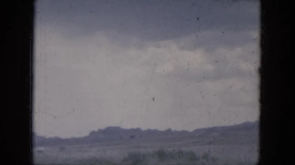 Late clip frame showing the Lake Canyon road landscape from a vehicle, with low hills, broad sky, and sparse vegetation visible through 8mm grain.