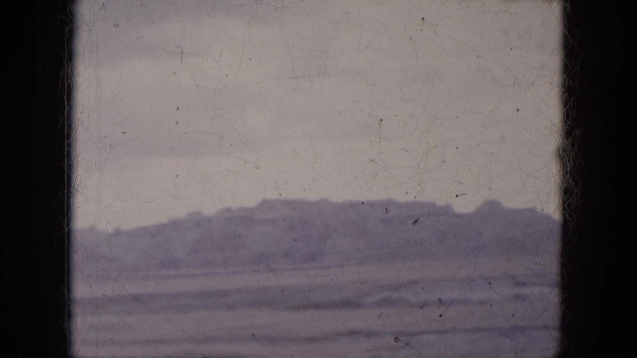 Muted 8mm frame of a desert ridge and broad flat terrain viewed from inside a moving car in Arizona.