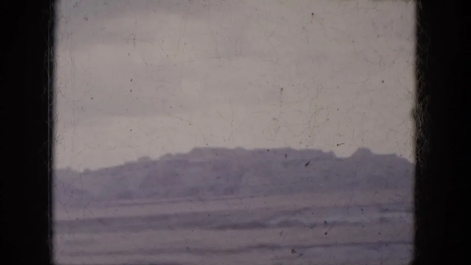 Muted 8mm frame of a desert ridge and broad flat terrain viewed from inside a moving car in Arizona.