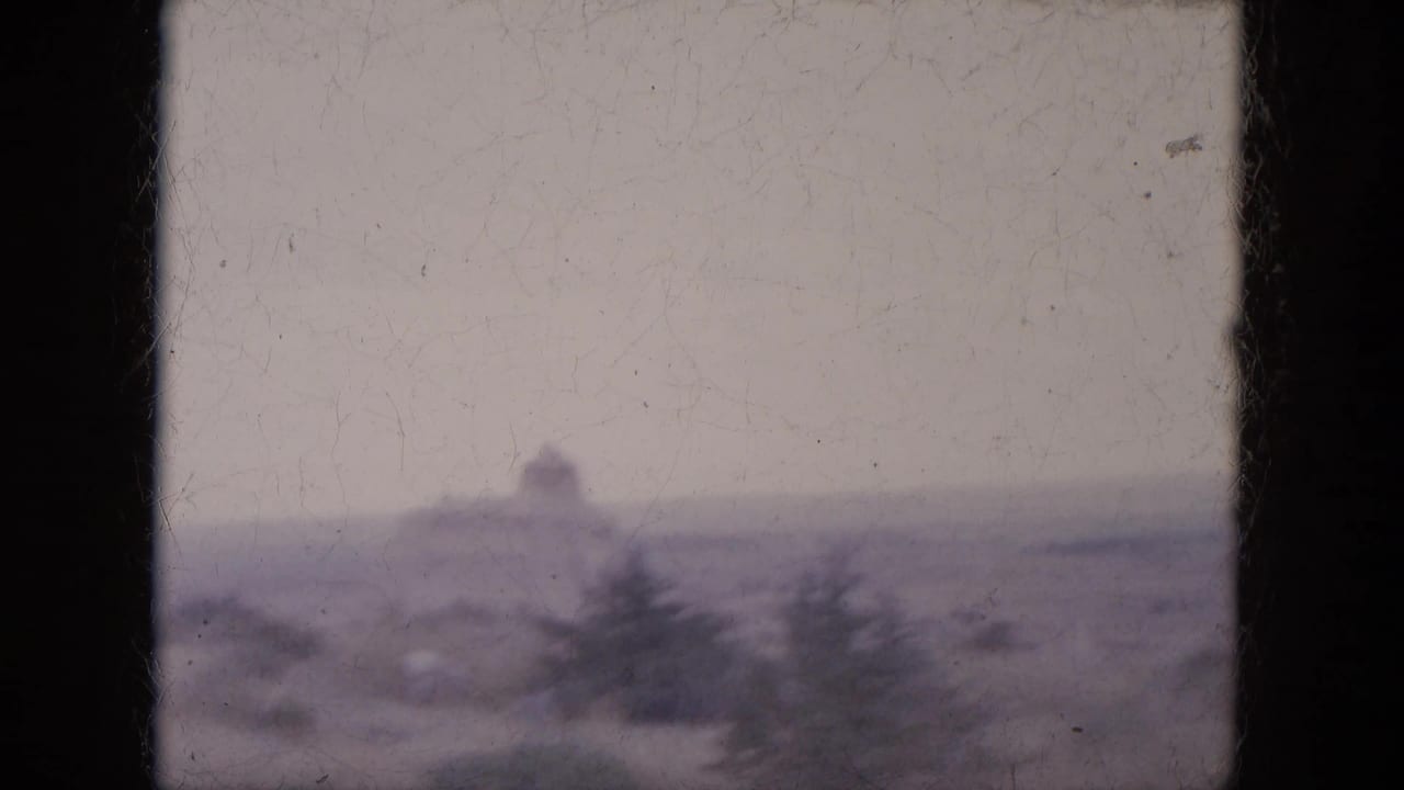 Hazy desert slope with a distant rock shape and sparse evergreen trees.