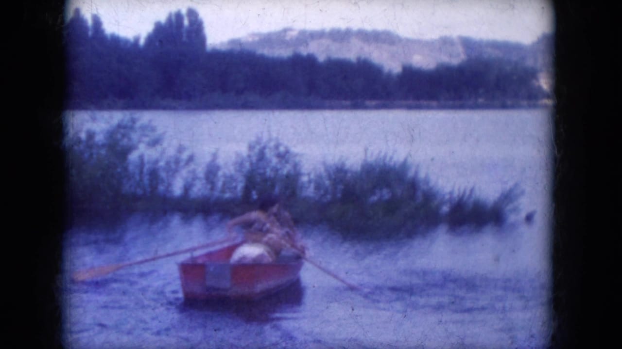 1969 8mm frame from Lake Canyon, Arizona, showing two people in a red rowboat drifting near reeds with hills beyond the lake.