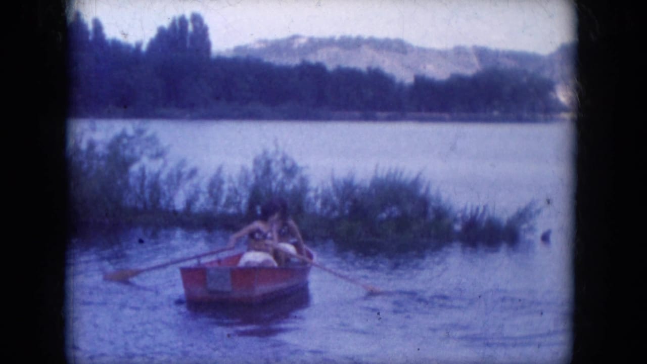 Two people sit low in a red rowboat on Lake Canyon, Arizona, with oars extended and reeds along the shoreline.