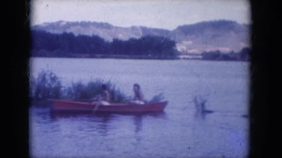 A side view shows the red rowboat with two people beside shoreline vegetation, with open water and wooded hills in the background.