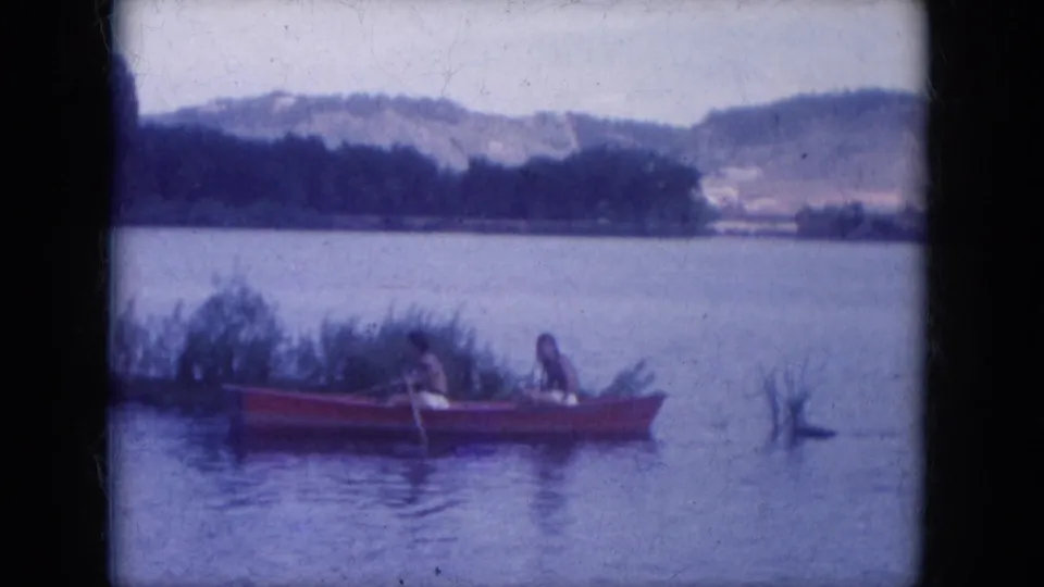 The red rowboat sits beside shoreline grasses on Lake Canyon, with two people aboard, a branch in the water, and hills beyond.