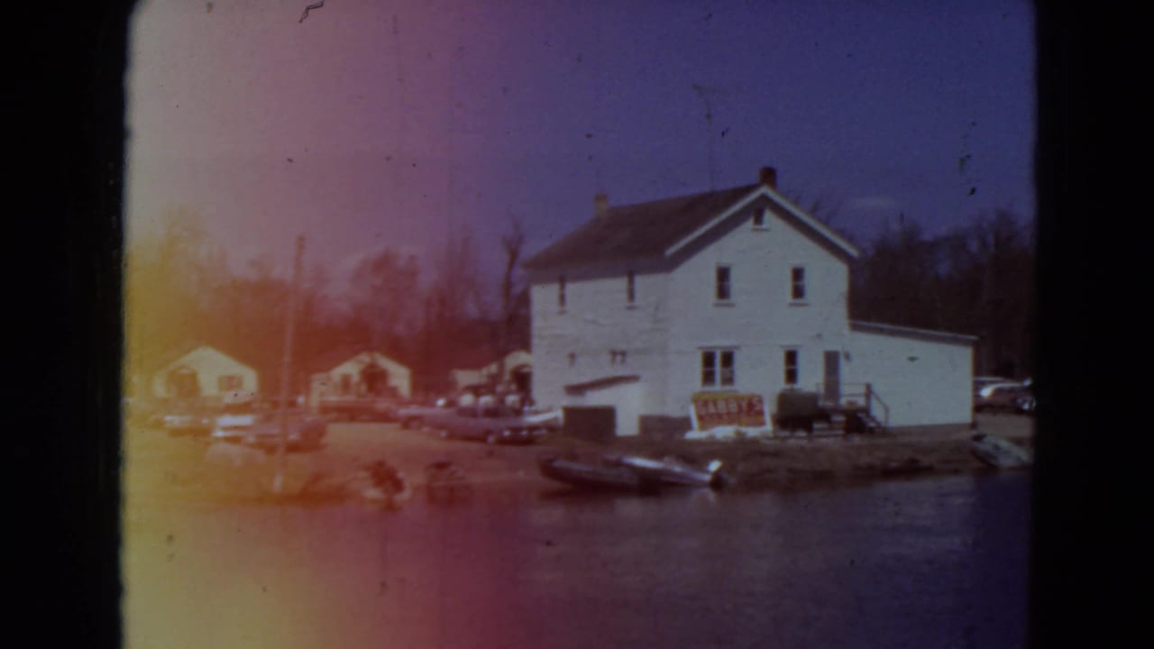 White two-story house by water with parked cars and small boats along the shore.