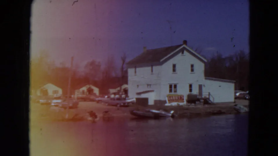 1969 Minnesota 8mm frame showing a white two-story waterfront building, parked cars, small boats, bare trees, and warm light leak at the left edge.
