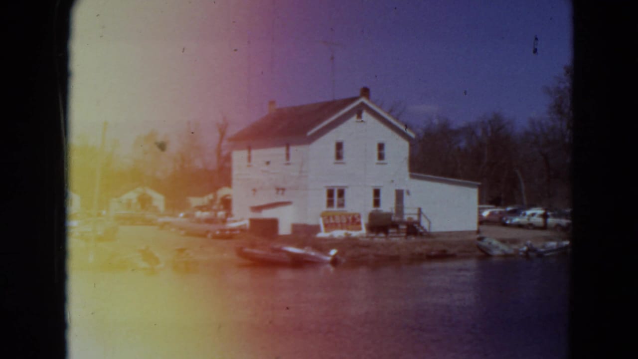 White house beside water with a row of cars and boats near the shoreline.