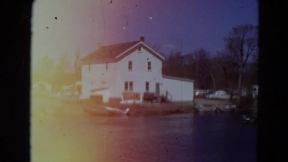 1969 Minnesota 8mm frame with the white waterfront house centered, boats at the shore, parked cars to the side, bare trees, and strong exposure bloom.