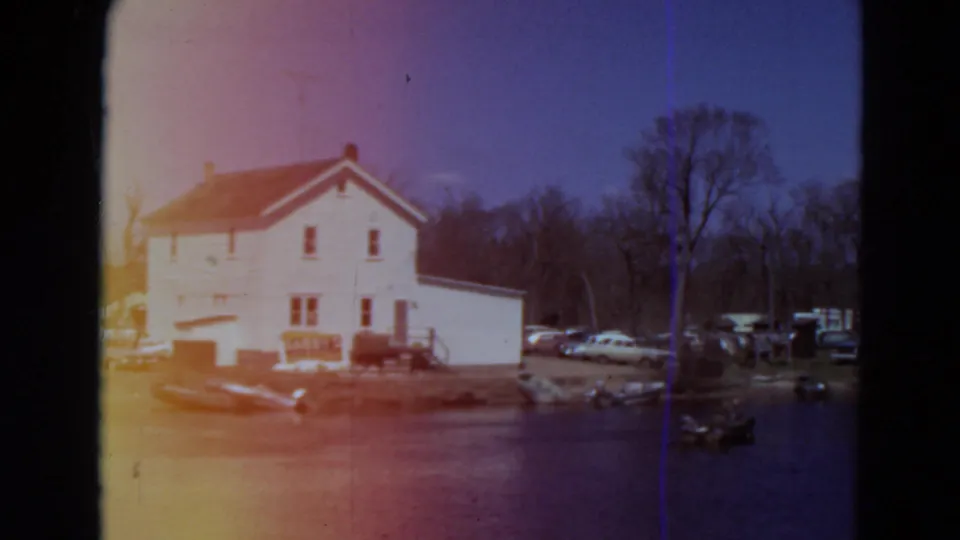 1969 Minnesota 8mm frame showing a white waterfront building, several cars, boats near the waterline, leafless trees, and a soft color-shifted scan.