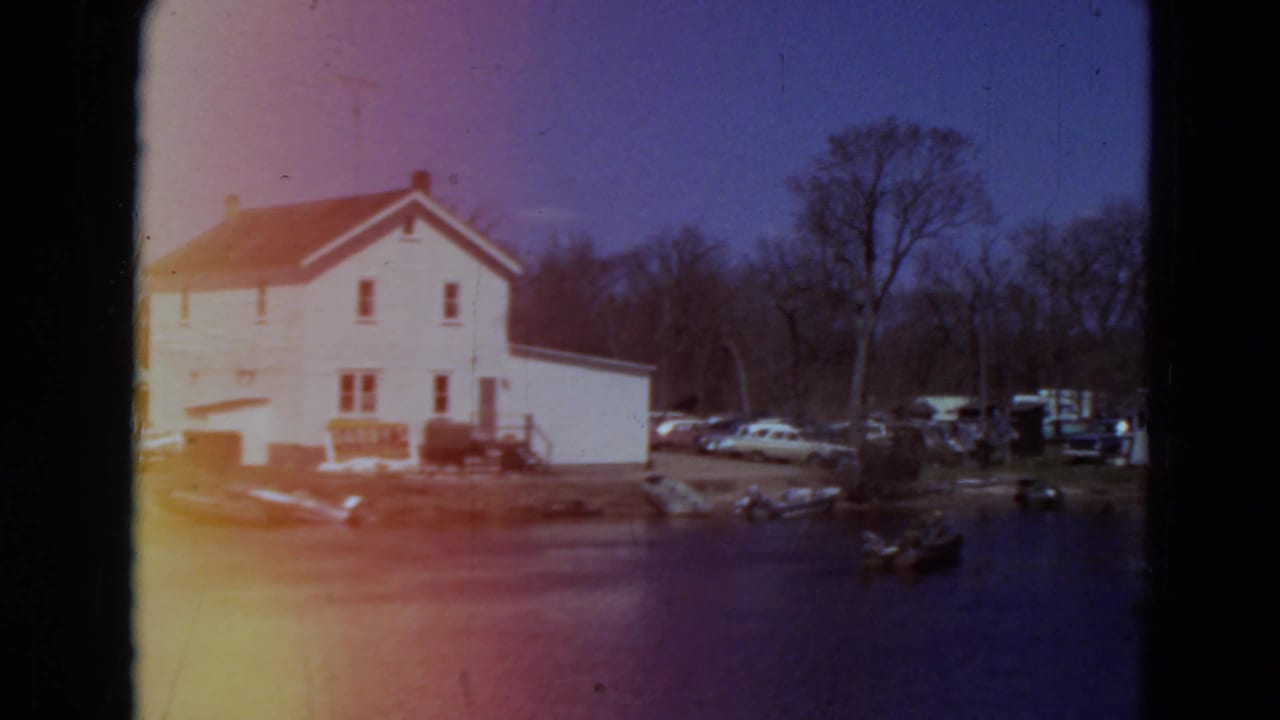 White lakeside house, parked cars, bare trees, and water with a small boat near shore.