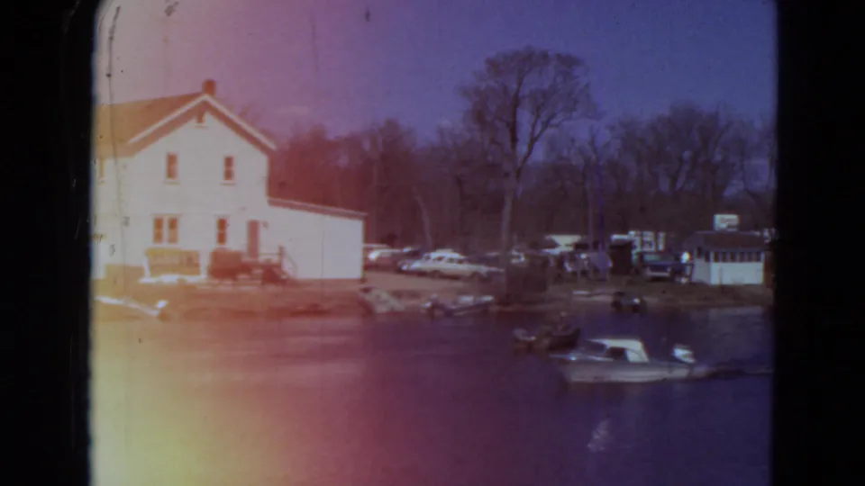 1969 Minnesota lakeside view with a speedboat crossing the water near a white house, parked cars, bare trees, and small shore structures.