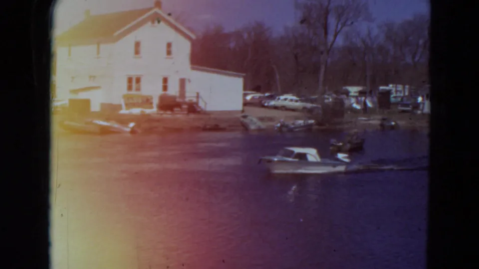 1969 Minnesota home-movie frame with a boat moving near the lower right, a white lakeside house, cars, docks, and leafless trees on shore.