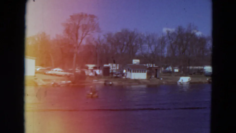 1969 Minnesota 8mm frame showing a wider shoreline with bare trees, small buildings, parked cars, open water, and a boat wake.