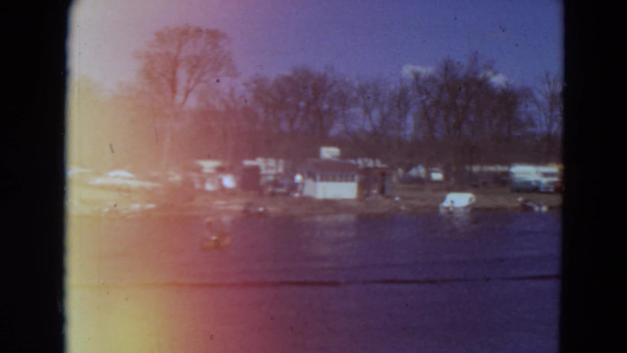 Lakeside campground with trailers, tents, bare trees, and water in the foreground.