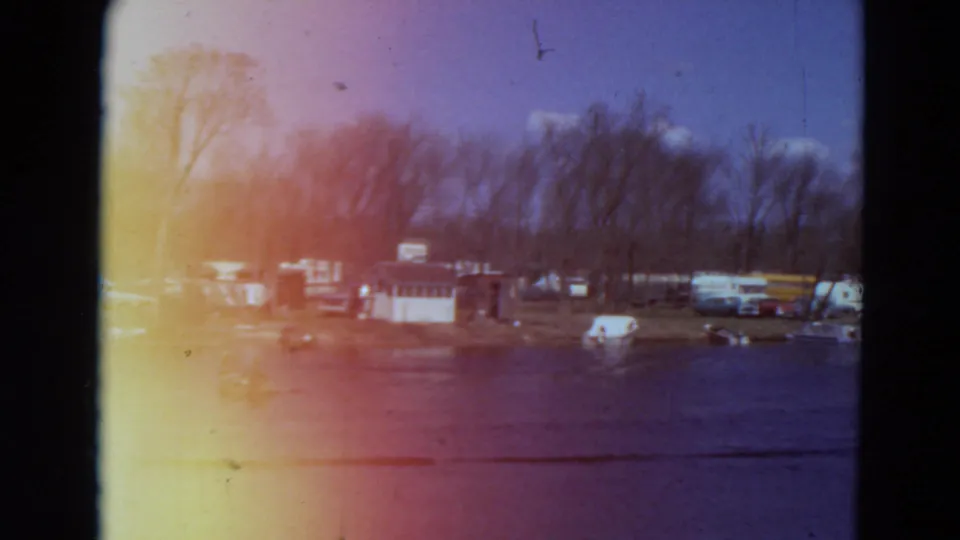 1969 Minnesota lakeside campground frame with parked trailers, tents, small boats, bare trees, and open water.