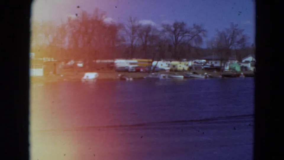 Wide 1969 Minnesota 8mm view of water facing a campground with trailers, tents, moored boats, and leafless trees.