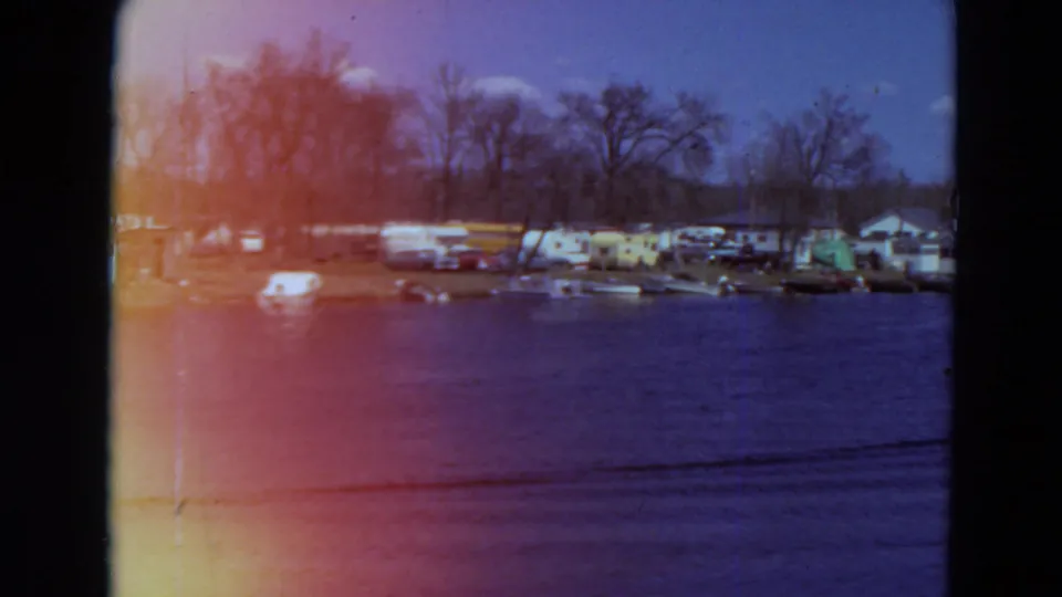 1969 Minnesota home-movie frame of a waterfront campground with trailers, tents, boats, bare trees, and visible 8mm color shift.