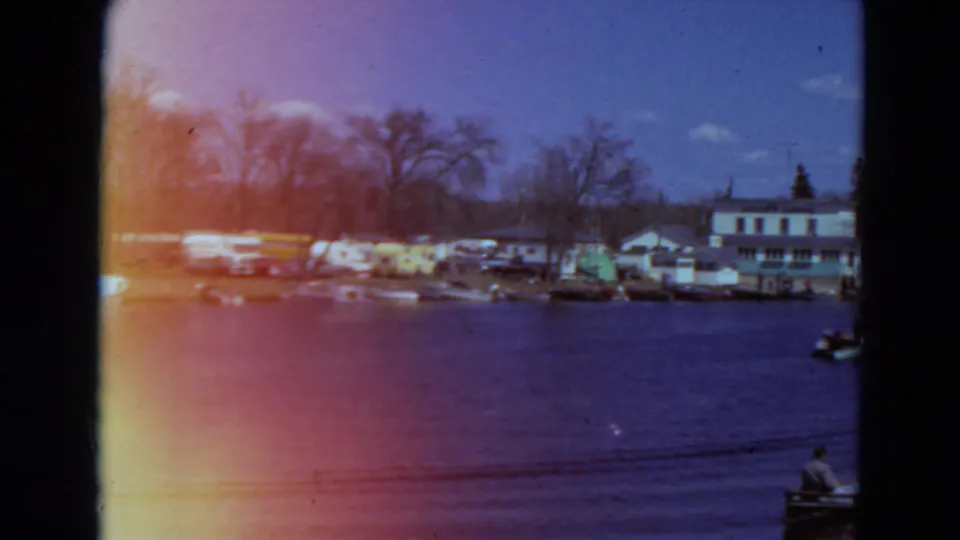 1969 Minnesota lakeside view with moored boats, shoreline buildings, bare trees, and a small craft with a person near the right edge.