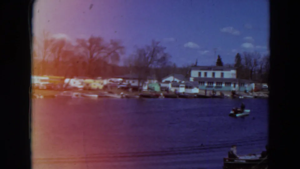 1969 Minnesota 8mm frame with a small motorboat crossing the lake in front of moored boats, houses, and bare shoreline trees.