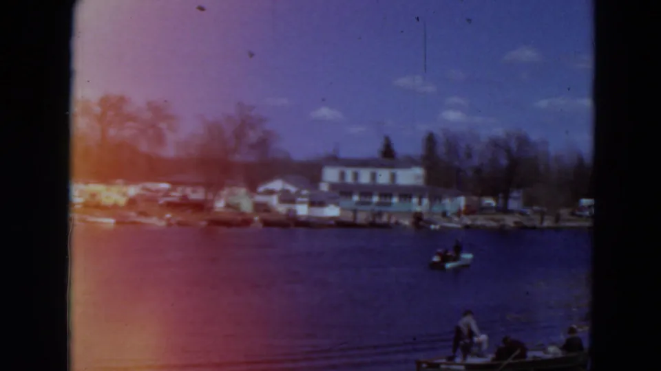 1969 Minnesota 8mm frame showing a lakeside building, parked cars, shoreline trees, and a small boat on calm water.