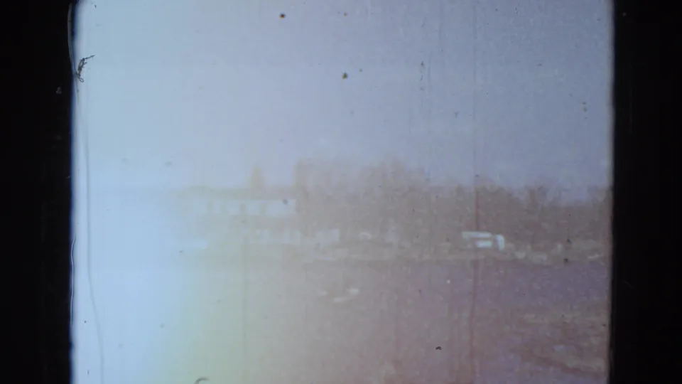 Overexposed 8mm frame from the Minnesota lake scene, with faint shoreline trees and vehicles or campers visible through color fading.