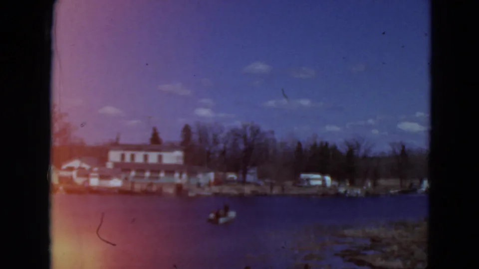 1969 Minnesota 8mm frame with a small boat on the lake, a shoreline building, parked cars, and trees under a bright sky.