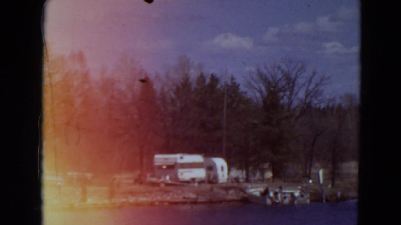 Lakeside campground with white trailers, parked cars, trees, and orange film flare.