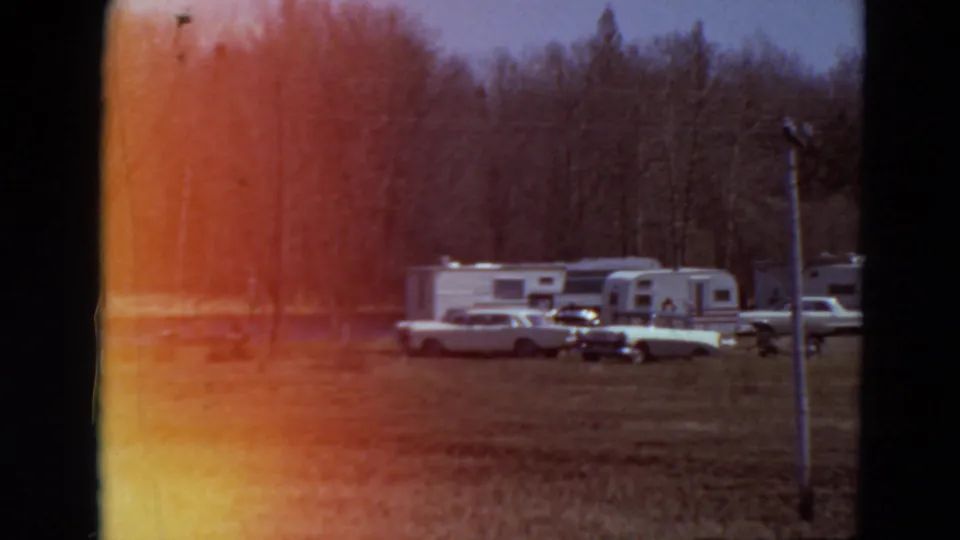 1969 Minnesota campground view with white travel trailers, classic cars, grass, bare trees, and warm film flare.