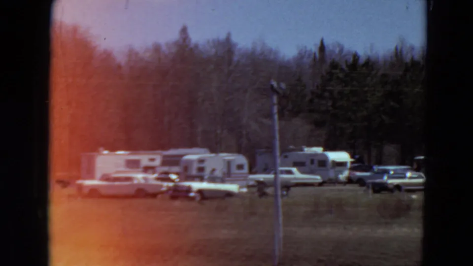 1969 Minnesota 8mm frame of trailers and mid-century cars in a grassy wooded campground, partly veiled by red flare.