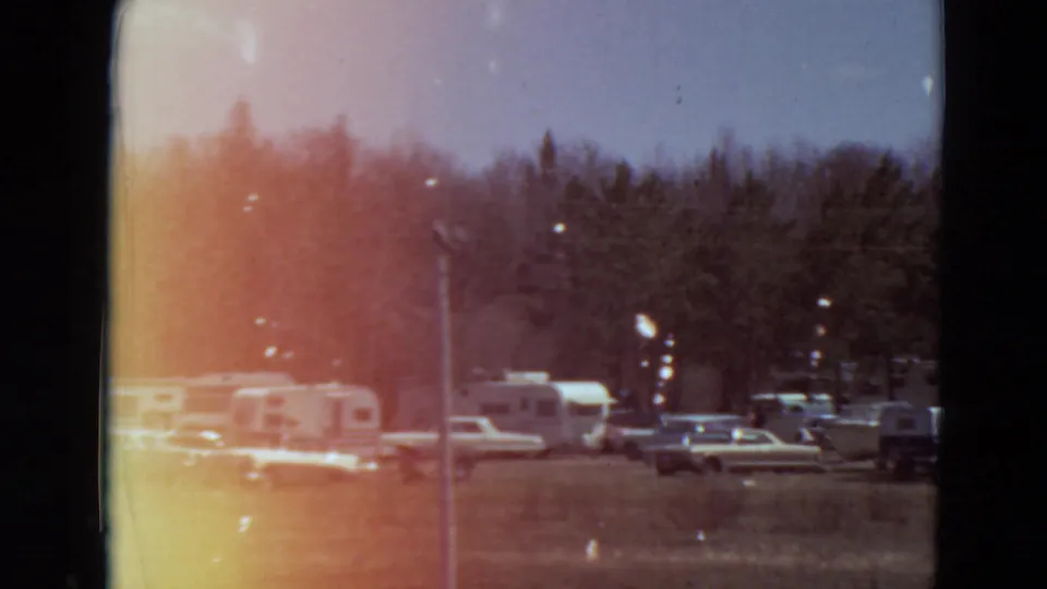 1969 Minnesota home-movie view of a wooded campground with trailers, cars, a pole, grass, and bright 8mm flare.