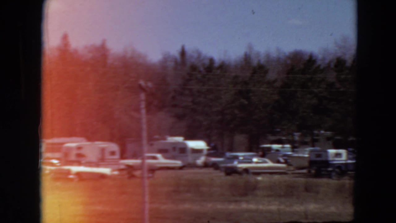 Campground with parked cars and trailers among trees, partly obscured by red film flare.