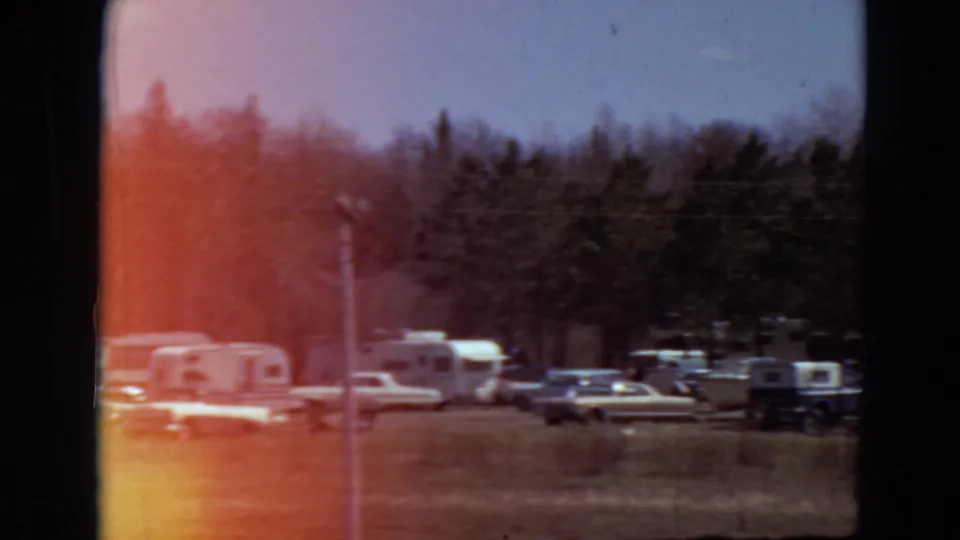 1969 Minnesota 8mm frame showing a distant campground with cars, trailers, trees, dry grass, and strong red-orange light leak.