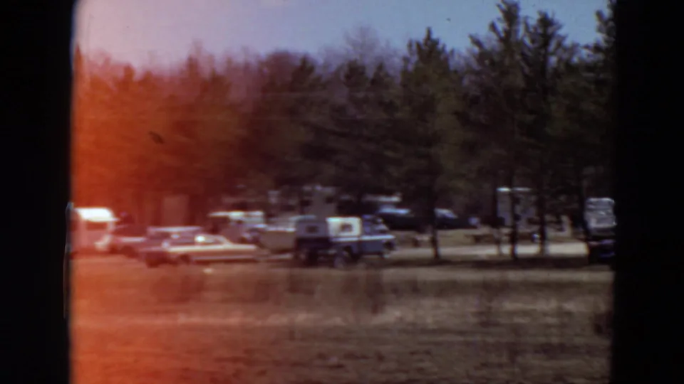 Distant 1969 Minnesota campground view with mid-century cars, trailers, pickup trucks, trees, grass, and soft 8mm exposure.