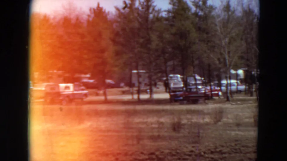 1969 Minnesota campground frame with parked vehicles, trailers, trees, dry ground, and a bright orange light leak from the film edge.
