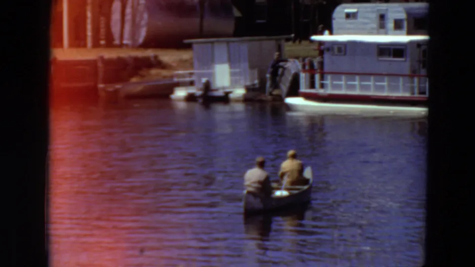1969 Minnesota lake scene with two adults canoeing near a dock, a houseboat-like vessel, and a small waterside structure.