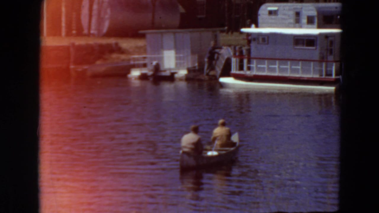 Two men in a canoe on a river near a docked houseboat and shoreline structure.
