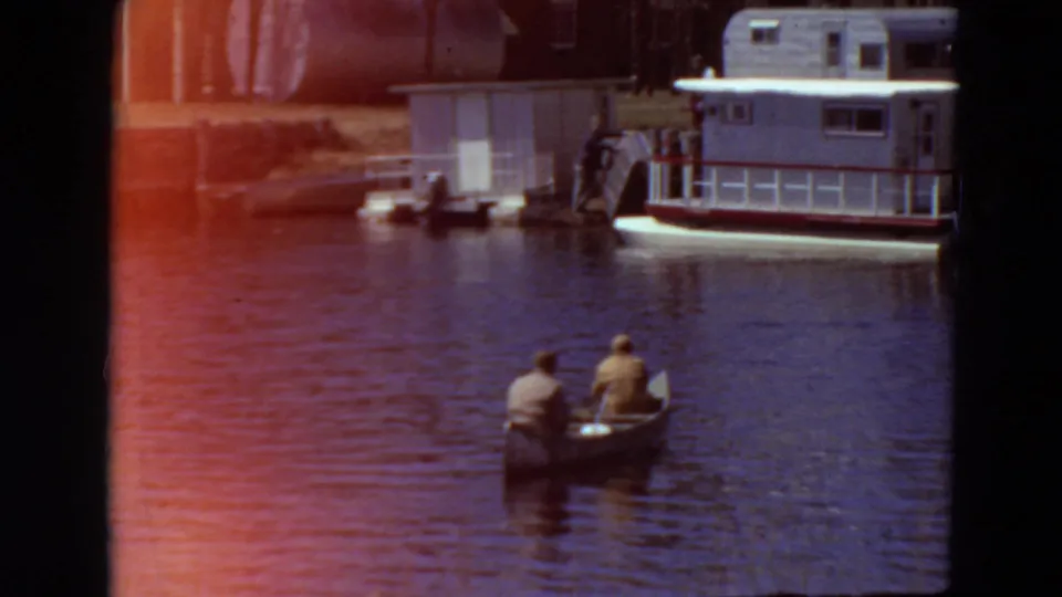 1969 Minnesota 8mm frame showing two men in a canoe on calm river water near a docked houseboat and small shore structure.